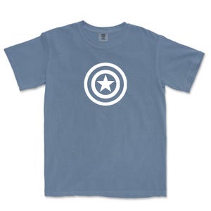 Puede incluir: Una camiseta azul de manga corta con un gr&aacute;fico blanco de una estrella dentro de un escudo circular. La camiseta est&aacute; hecha de un material suave y tiene cuello redondo. El dise&ntilde;o est&aacute; centrado en la parte delantera de la camiseta.