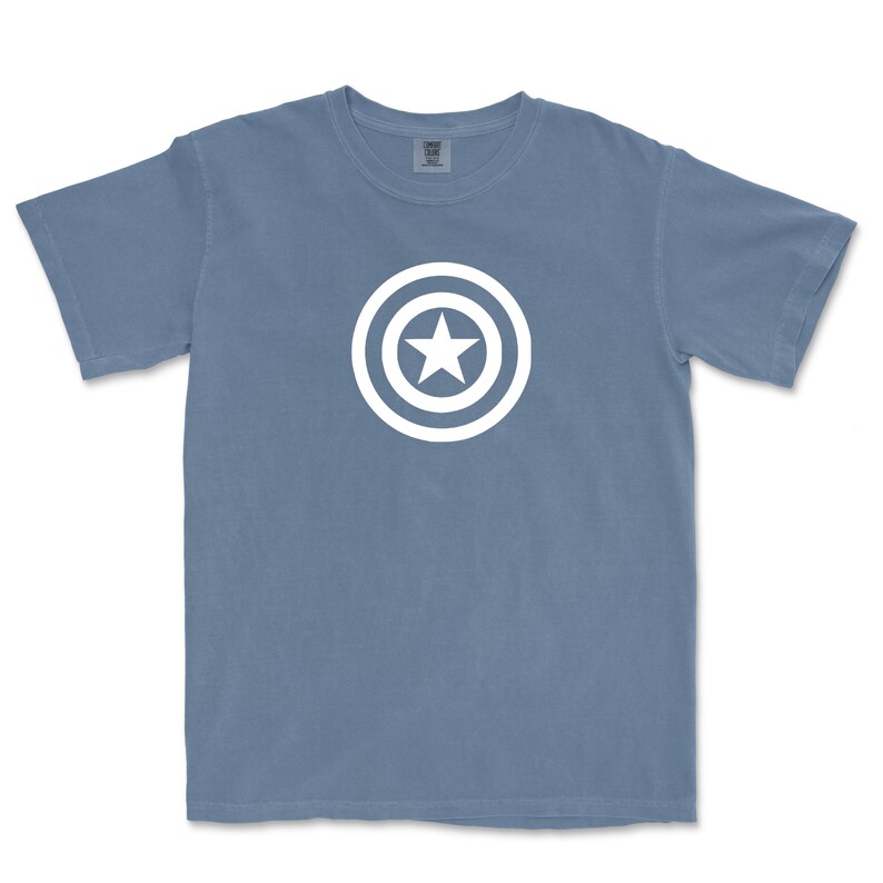 Puede incluir: Una camiseta azul de manga corta con un gr&aacute;fico blanco de una estrella dentro de un escudo circular. La camiseta est&aacute; hecha de un material suave y tiene cuello redondo. El dise&ntilde;o est&aacute; centrado en la parte delantera de la camiseta.
