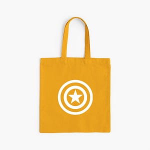 K&ouml;nnte beinhalten: Senfgelbe Tragetasche mit wei&szlig;em Captain America Schild-Design. Die Tasche hat eine rechteckige Form mit passenden Griffen. Die Grafik zeigt einen wei&szlig;en Stern in einem wei&szlig;en Kreis mit wei&szlig;em Rand.