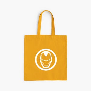 May include: Mustard yellow tote bag featuring a white Iron Man helmet logo within a white circle. The bag has matching yellow handles and is made of a sturdy fabric. Perfect for carrying books, groceries, or everyday essentials.