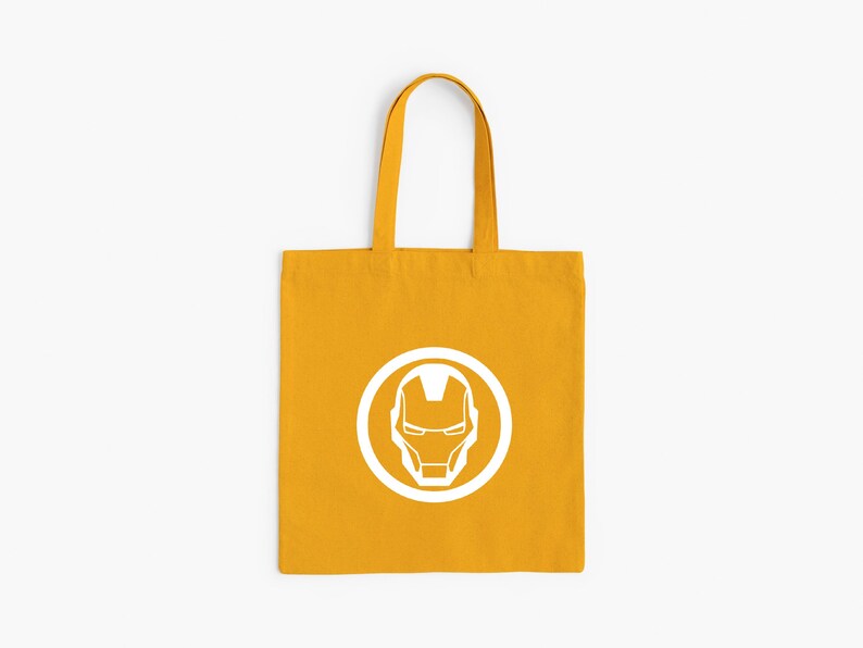 May include: Mustard yellow tote bag featuring a white Iron Man helmet logo within a white circle. The bag has matching yellow handles and is made of a sturdy fabric. Perfect for carrying books, groceries, or everyday essentials.