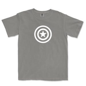 Puede incluir: Una camiseta gris con un dise&ntilde;o gr&aacute;fico blanco de una estrella dentro de un escudo circular. La camiseta tiene mangas cortas y cuello redondo. El dise&ntilde;o est&aacute; centrado en el pecho.