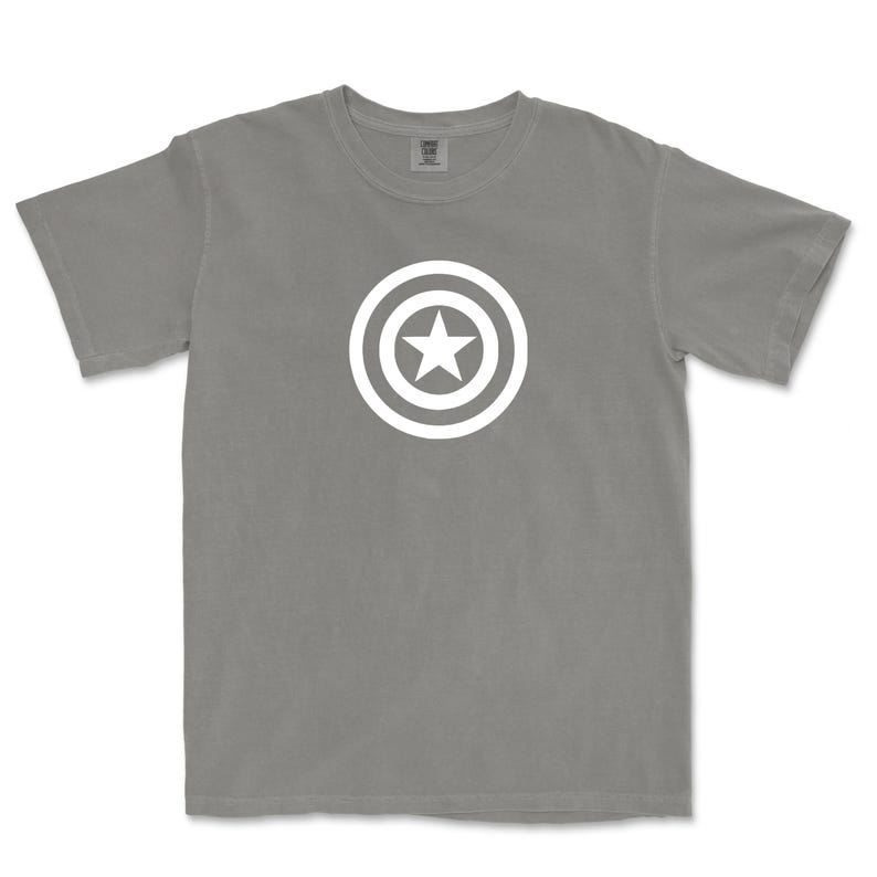 Puede incluir: Una camiseta gris con un dise&ntilde;o gr&aacute;fico blanco de una estrella dentro de un escudo circular. La camiseta tiene mangas cortas y cuello redondo. El dise&ntilde;o est&aacute; centrado en el pecho.