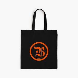 Op de afbeelding: Zwarte draagtas met een oranje cirkelvormig ontwerp met een gestileerde letter "B". De tas heeft een eenvoudig ontwerp met een rechthoekige body en twee zwarte handvatten om te dragen.