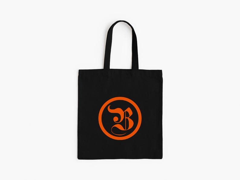 Op de afbeelding: Zwarte draagtas met een oranje cirkelvormig ontwerp met een gestileerde letter "B". De tas heeft een eenvoudig ontwerp met een rechthoekige body en twee zwarte handvatten om te dragen.