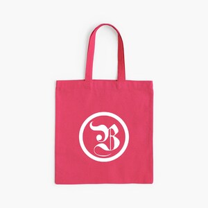 Op de afbeelding: Een felroze draagtas met een wit cirkelvormig logo met een gestileerde letter "B". De tas heeft een eenvoudig ontwerp met bijpassende roze handvatten, geschikt voor het dragen van boeken, boodschappen of persoonlijke spullen.