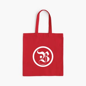 Op de afbeelding: Een rode draagtas met witte handvatten. De tas heeft een wit cirkelvormig ontwerp met een gestileerde letter "B" erin. De tas is gemaakt van een stevige stof en is geschikt voor het dragen van diverse items.