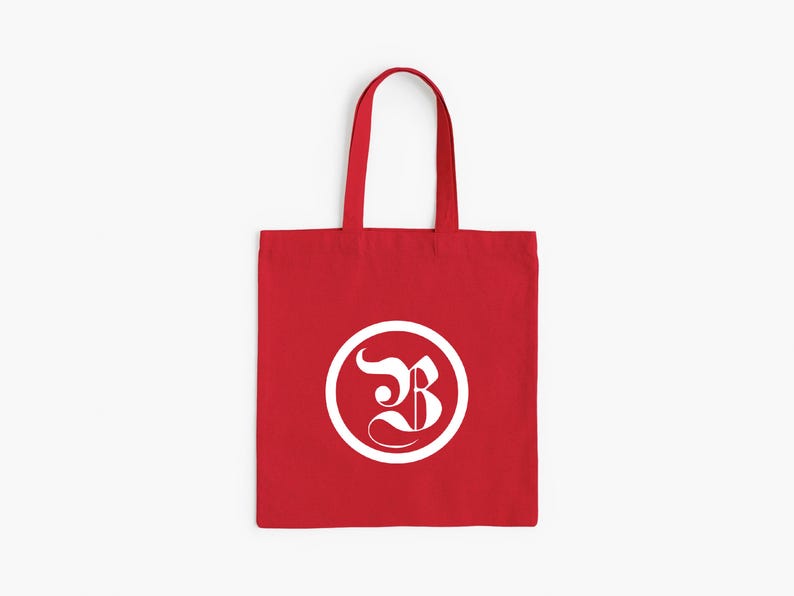 Op de afbeelding: Een rode draagtas met witte handvatten. De tas heeft een wit cirkelvormig ontwerp met een gestileerde letter "B" erin. De tas is gemaakt van een stevige stof en is geschikt voor het dragen van diverse items.