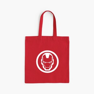 May include: Red tote bag featuring a white Iron Man helmet design inside a white circle. The bag has a rectangular shape with a handle for carrying. The design is centered on the front of the bag.