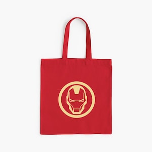 May include: Red tote bag featuring a gold Iron Man logo within a gold circle. The bag has a rectangular shape with a handle for carrying. The design is centered on the front of the bag.
