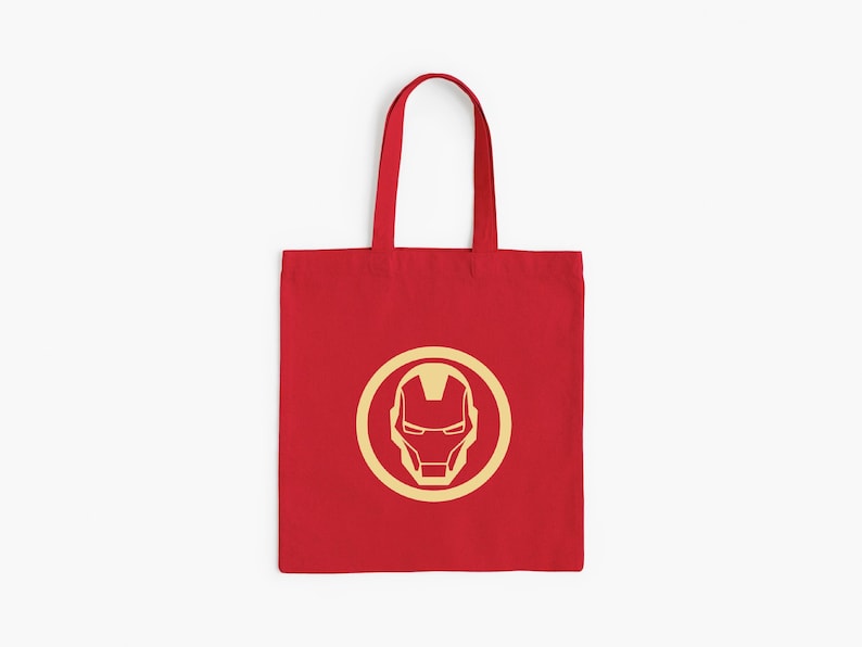 May include: Red tote bag featuring a gold Iron Man logo within a gold circle. The bag has a rectangular shape with a handle for carrying. The design is centered on the front of the bag.