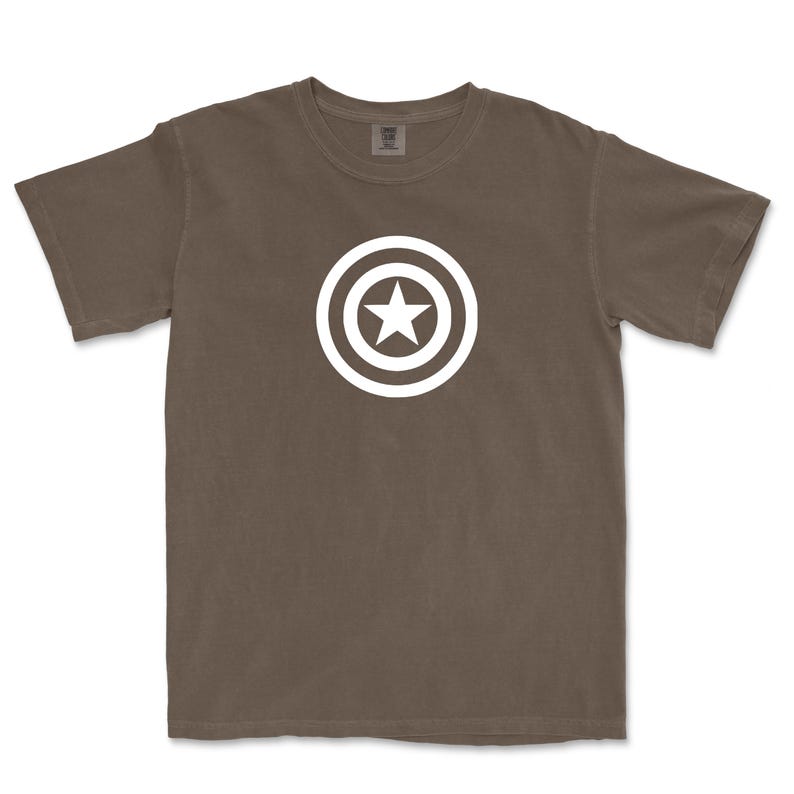 Puede incluir: Camiseta marr&oacute;n con un dise&ntilde;o gr&aacute;fico blanco de una estrella dentro de un c&iacute;rculo, que recuerda a un escudo. La camiseta es de material suave y tiene mangas cortas. El dise&ntilde;o est&aacute; centrado en el pecho.