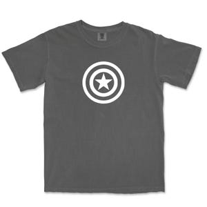 Puede incluir: Una camiseta gris oscuro con un dise&ntilde;o gr&aacute;fico blanco de una estrella dentro de un escudo circular. La camiseta tiene cuello redondo y mangas cortas. El dise&ntilde;o est&aacute; centrado en el pecho.