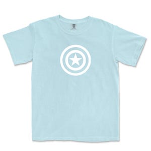 Puede incluir: Camiseta azul claro con un dise&ntilde;o gr&aacute;fico blanco de una estrella dentro de un escudo circular. La camiseta de manga corta tiene cuello redondo. El gr&aacute;fico est&aacute; centrado en la parte delantera.