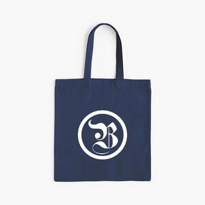 Op de afbeelding: Marineblauwe draagtas met wit cirkelvormig logo. Het logo toont de gestileerde witte letter "B" in een witte cirkel. De tas heeft een eenvoudige rechthoekige vorm en twee handvatten.