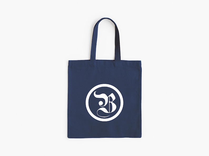 Op de afbeelding: Marineblauwe draagtas met wit cirkelvormig logo. Het logo toont de gestileerde witte letter "B" in een witte cirkel. De tas heeft een eenvoudige rechthoekige vorm en twee handvatten.