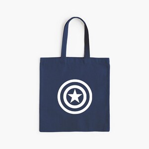 K&ouml;nnte beinhalten: Marineblaue Tragetasche mit einem wei&szlig;en Stern-in-Kreis-Grafik. Die Tasche hat eine rechteckige Form mit einem Griff zum Tragen. Das Design befindet sich mittig auf der Vorderseite der Tasche.