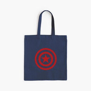 K&ouml;nnte beinhalten: Marineblaue Tragetasche mit rotem Schild-Design und Stern in der Mitte. Die Tasche hat eine rechteckige Form und einen Griff. Das Design ist auf der Vorderseite der Tasche zentriert. Geeignet f&uuml;r den Transport von Gegenst&auml;nden.