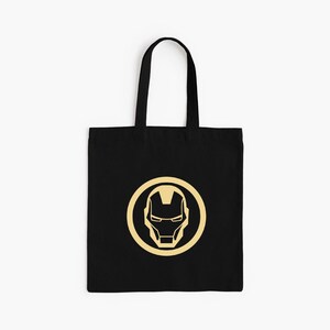 May include: Black tote bag featuring a gold Iron Man mask design within a gold circle. The bag has black handles and is made of a durable fabric. A stylish and practical accessory for carrying everyday items.