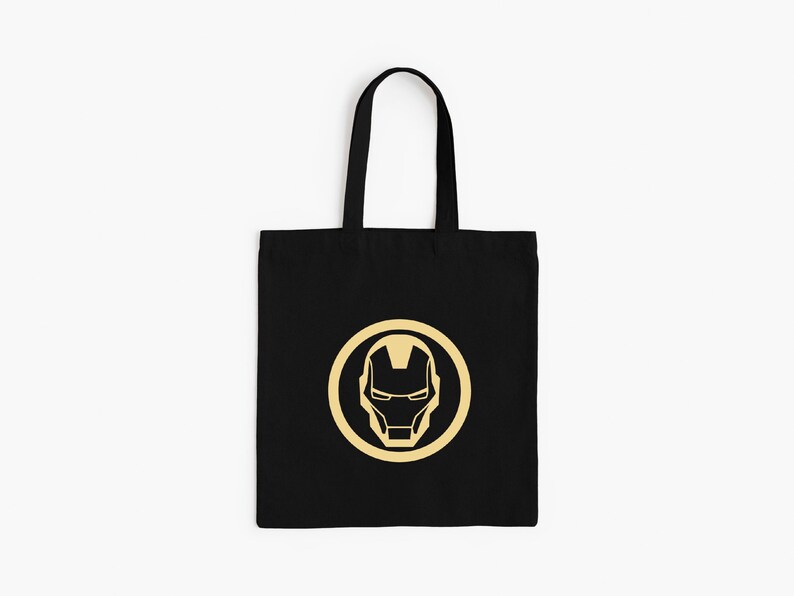 May include: Black tote bag featuring a gold Iron Man mask design within a gold circle. The bag has black handles and is made of a durable fabric. A stylish and practical accessory for carrying everyday items.