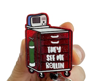 May include: Red glitter medical cart with a white "They See Me Rollin'" text. The cart has a small monitor, a green oxygen tank, and four wheels. The cart is attached to a retractable badge reel.