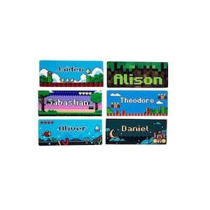 May include: Six rectangular name tags with pixelated video game-themed designs. Each tag features a different name: Jaiden, Alison, Sabastian, Theodore, Oliver, and Daniel. The tags have colourful backgrounds and pixel art graphics.