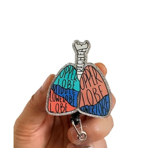 May include: A silver glitter badge reel with a colorful anatomical illustration of a lung with the words "upper lobe", "middle lobe", "lower lobe", and "trachea" printed on it.