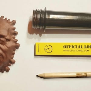 Könnte beinhalten: Ein braunes, 3D-gedrucktes Gesicht mit einem Schmelzeffekt, ein gelbes Logbuch mit dem Text "OFFICIAL LOGBOOK www.geocaching.com", ein schwarzes Metallrohr und ein Bleistift aus Holz.