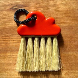 May include: A small, decorative brush with a bright orange cloud-shaped handle and natural-colored bristles. The handle has a black cord loop for hanging. The brush is set against a wooden background.