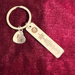 May include: Silver keychain with a heart-shaped charm and a rectangular tag. The heart charm has the letter "N" engraved on it. The rectangular tag features a music-themed design and a barcode-like pattern.