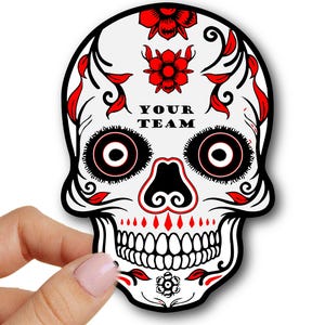 Puede incluir: Pegatina de calavera de azúcar blanca con detalles negros y rojos. La calavera tiene ojos grandes con pupilas negras y contornos rojos. La calavera está decorada con flores rojas y remolinos negros. El texto "YOUR TEAM" está impreso en la frente de la calavera.