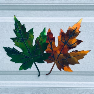 May include: Two metallic maple leaves, one green and one orange, are attached to a white wall. The leaves are cut out and have a shiny, reflective finish.