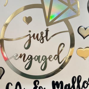 May include: A white decal sheet featuring a large gold ring outline with a holographic diamond, the text "just engaged" in gold script, and the black text "cg & mallory" below. The design includes various gold and black heart-shaped stickers.