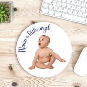 May include: A white round mousepad with a baby sitting on it. The baby is wearing a diaper and has a surprised expression on their face. The text "Mama's little angel" is written in blue cursive around the baby.