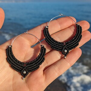 Könnte beinhalten: Ein Paar schwarze Makramee-Creolen mit silberfarbenen Akzenten. Die Ohrringe haben eine halbmondförmige Form mit einem Perlendesign und einer kleinen silbernen Perle unten. Sie werden vor dem Hintergrund des Meeres präsentiert.