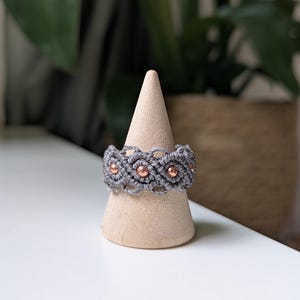 May include: A grey macrame ring featuring a woven cord pattern with small, round, metallic copper-colored beads integrated into the design. The ring is displayed on a smooth, light-colored wooden cone-shaped stand for clear visibility.