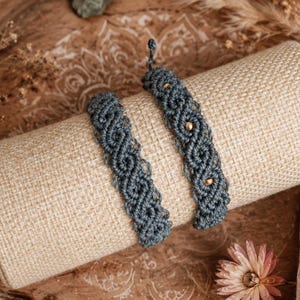 Könnte beinhalten: Zwei Makramee-Armbänder in einem gedämpften Blau-Grau, präsentiert auf einem beigen zylindrischen Ständer. Ein Armband hat goldfarbene Perlen. Die Armbänder werden auf einem Holztablett mit dekorativen Elementen wie Trockenblumen und einem kleinen grünen Stein präsentiert.