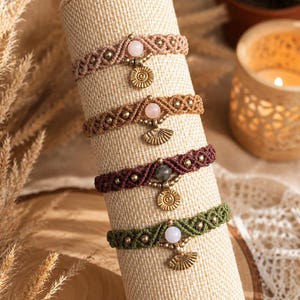 May include: Four macrame bracelets in beige, brown, dark red, and olive green. Each bracelet features a central round gemstone and a dangling antique gold-toned metal charm in either a circular sunburst or a fan shape, secured by woven cord.