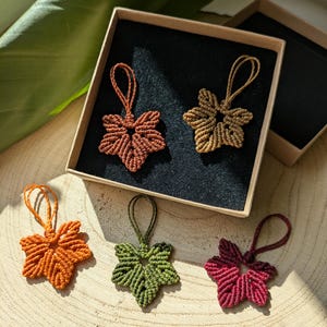 May include: Five macrame leaf ornaments in various colors including rust, tan, orange, olive green, and deep red. Each leaf features a knotted cord loop at the top for hanging. Two ornaments are inside a square cardboard box with black lining.