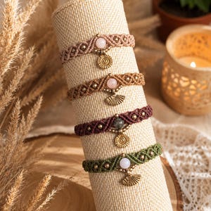 May include: Five macrame bracelets in shades of pink, brown, and green are displayed on a beige cylinder. Each bracelet features a gemstone and a gold-coloured charm. The bracelets are adorned with small, gold-coloured beads.