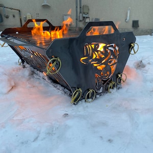 May include: A black metal fire pit with a cut-out design of a fish. The fire pit is burning with orange flames and is sitting on a snowy surface.