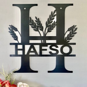 May include: Black metal monogram letter H with the name "HAFSO" written below. The letter is decorated with wheat stalks.