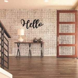 May include: A black metal "Hello" sign hangs on a wall with a textured pattern. A wooden door with glass panels is to the right of the sign. A console table with two lamps sits in front of the wall.