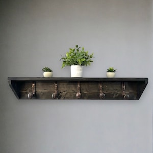 Peut inclure: Une étagère en bois teinté foncé avec cinq crochets en métal, supportant trois petites plantes en pot. L'étagère est fixée sur un mur gris, avec une porte blanche et un petit banc noir en arrière-plan. Solution de rangement fonctionnelle et décorative.