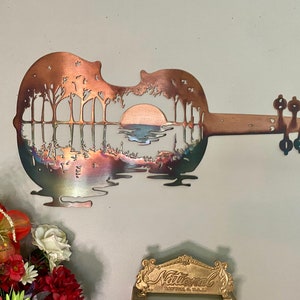 May include: A metal wall art sculpture of a guitar with a sunset and trees reflected in the water inside the body of the guitar. The guitar is made of copper and has a rustic finish.