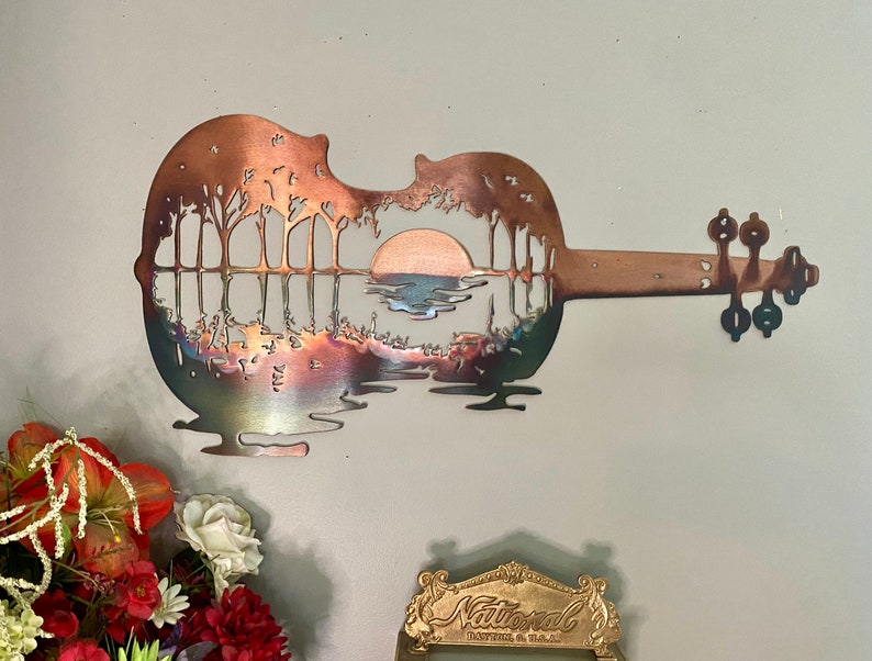 May include: A metal wall art sculpture of a guitar with a sunset and trees reflected in the water inside the body of the guitar. The guitar is made of copper and has a rustic finish.