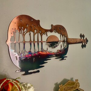 May include: A metal wall sculpture of a violin with a sunset scene and trees silhouetted against the horizon. The sculpture is made of copper and has a rainbow iridescent finish.