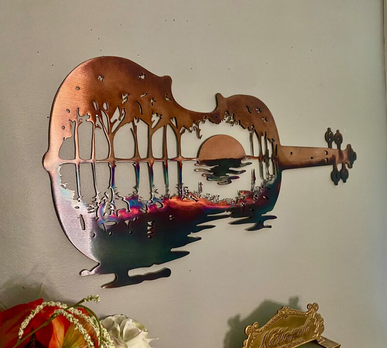 May include: A metal wall sculpture of a violin with a sunset scene and trees silhouetted against the horizon. The sculpture is made of copper and has a rainbow iridescent finish.