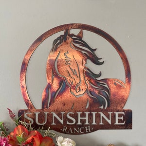 Peut inclure: Une enseigne en métal avec une silhouette de tête de cheval dans un cercle. L'enseigne est peinte en cuivre avec une finition rustique. Le texte "SUNSHINE RANCH" est situé sous la tête de cheval.