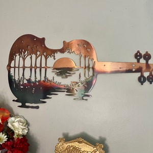 May include: A metal wall art sculpture of a guitar with a sunset scene cut out of the body. The guitar is a copper color and the scene is a silhouette of trees and a lake with a setting sun.